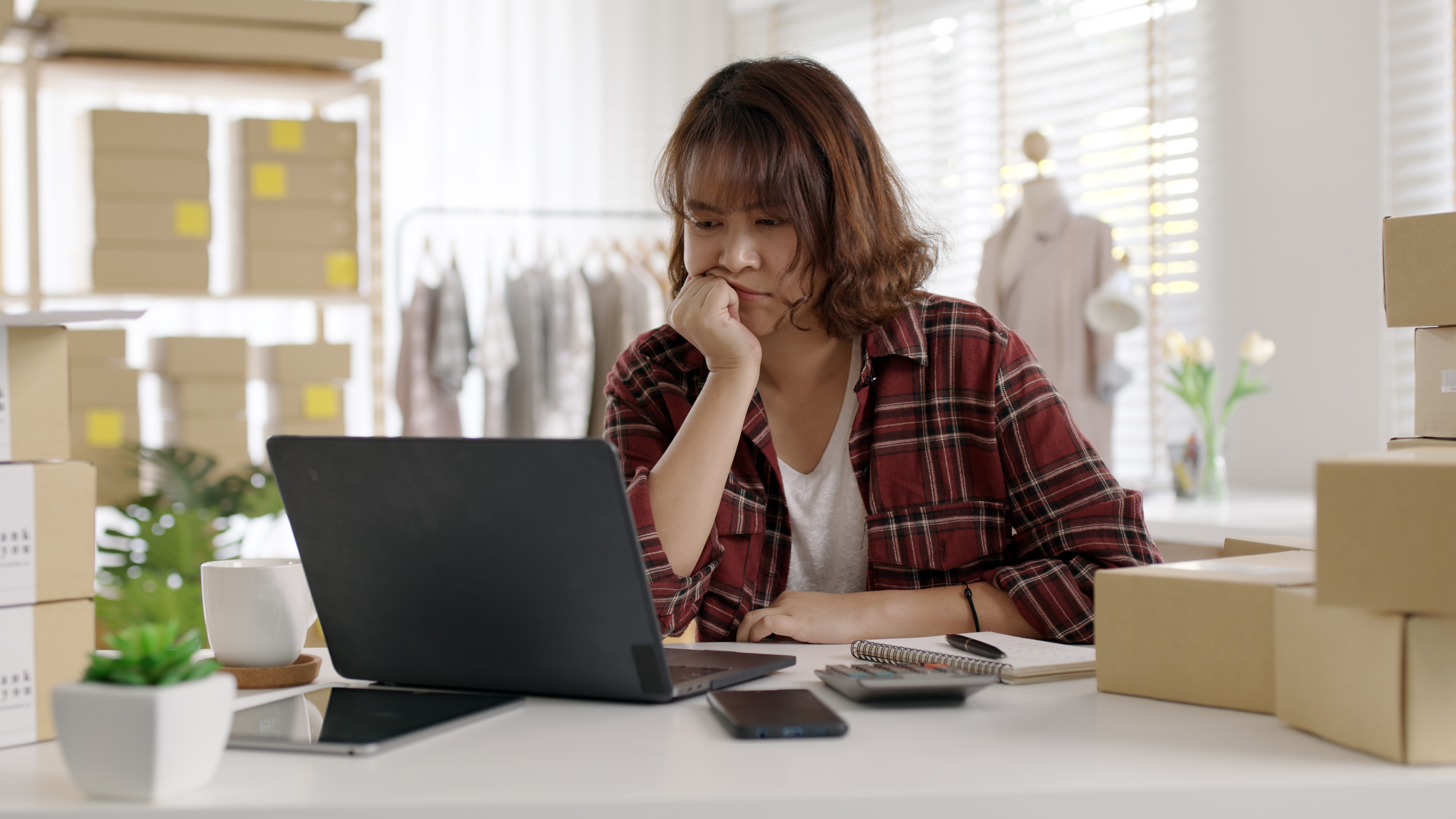 A woman working on a laptop at a desk, appearing focused. She is wearing a red plaid shirt and surrounded by business tools including a smartphone, calculator, and cardboard box, suggesting she may be running an online retail or fashion business.
