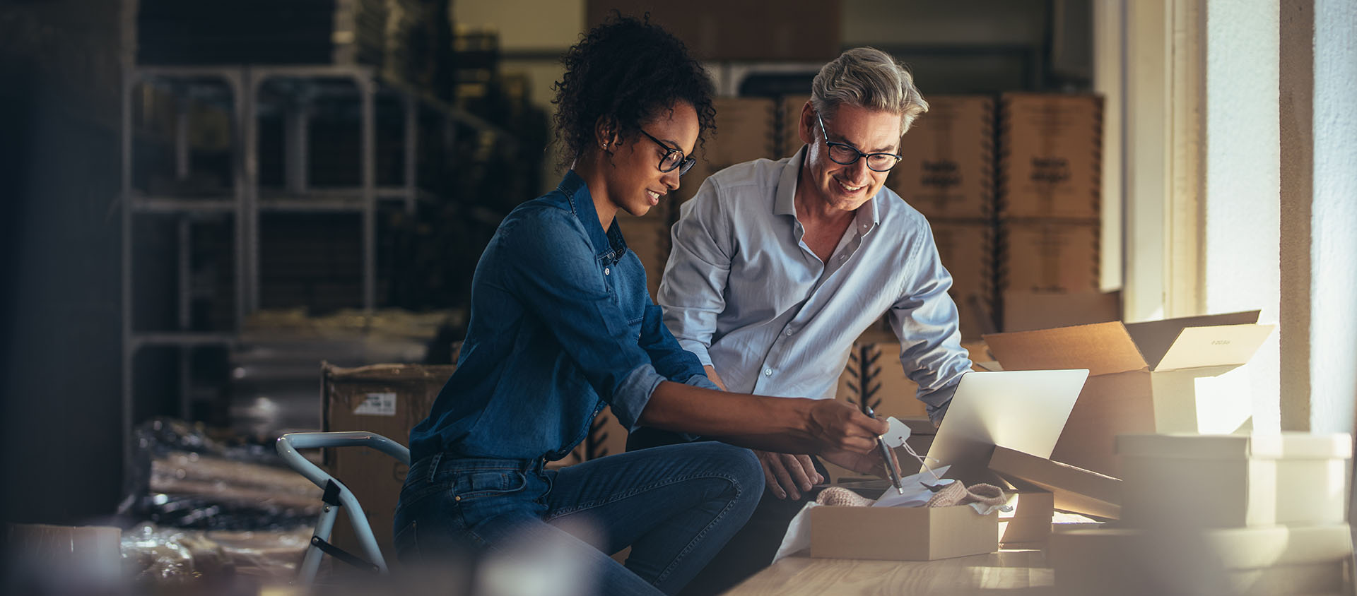 A woman and a man working together in a warehouse, reviewing items on a table with boxes in the background.